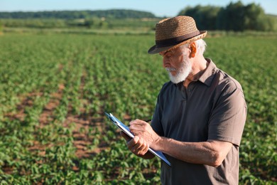 Photo of Senior farmer with clipboard in field of unripe soy outdoors, space for text