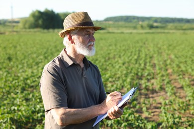 Photo of Senior farmer with clipboard in field of unripe soy outdoors, space for text