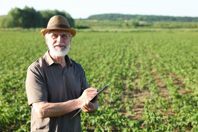 Photo of Senior farmer with clipboard in field of unripe soy outdoors, space for text