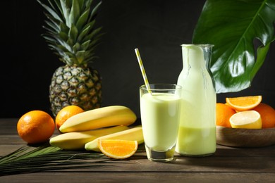 Photo of Superfood. Refreshing smoothie in bottle, glass, palm leaf and ingredients on wooden table against black background, closeup