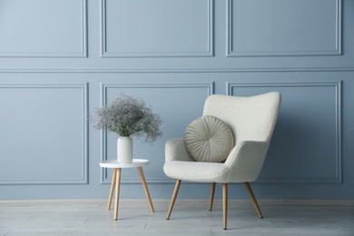 Photo of Comfortable armchair and vase with flowers near light blue wall indoors