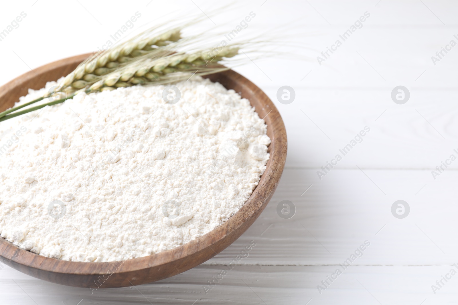 Photo of Flour and green wheat spikes in bowl on white wooden table, closeup. Space for text