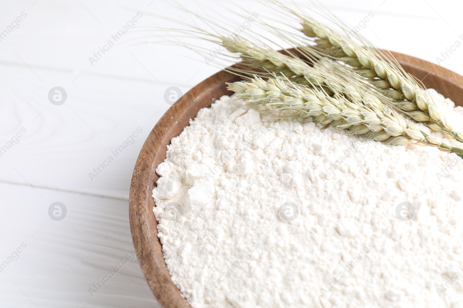 Photo of Flour and green wheat spikes in bowl on white wooden table, closeup