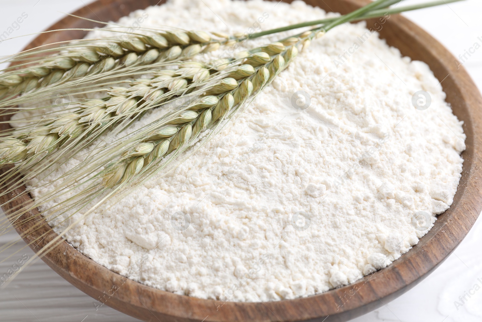 Photo of Flour and green wheat spikes in bowl on white wooden table, closeup