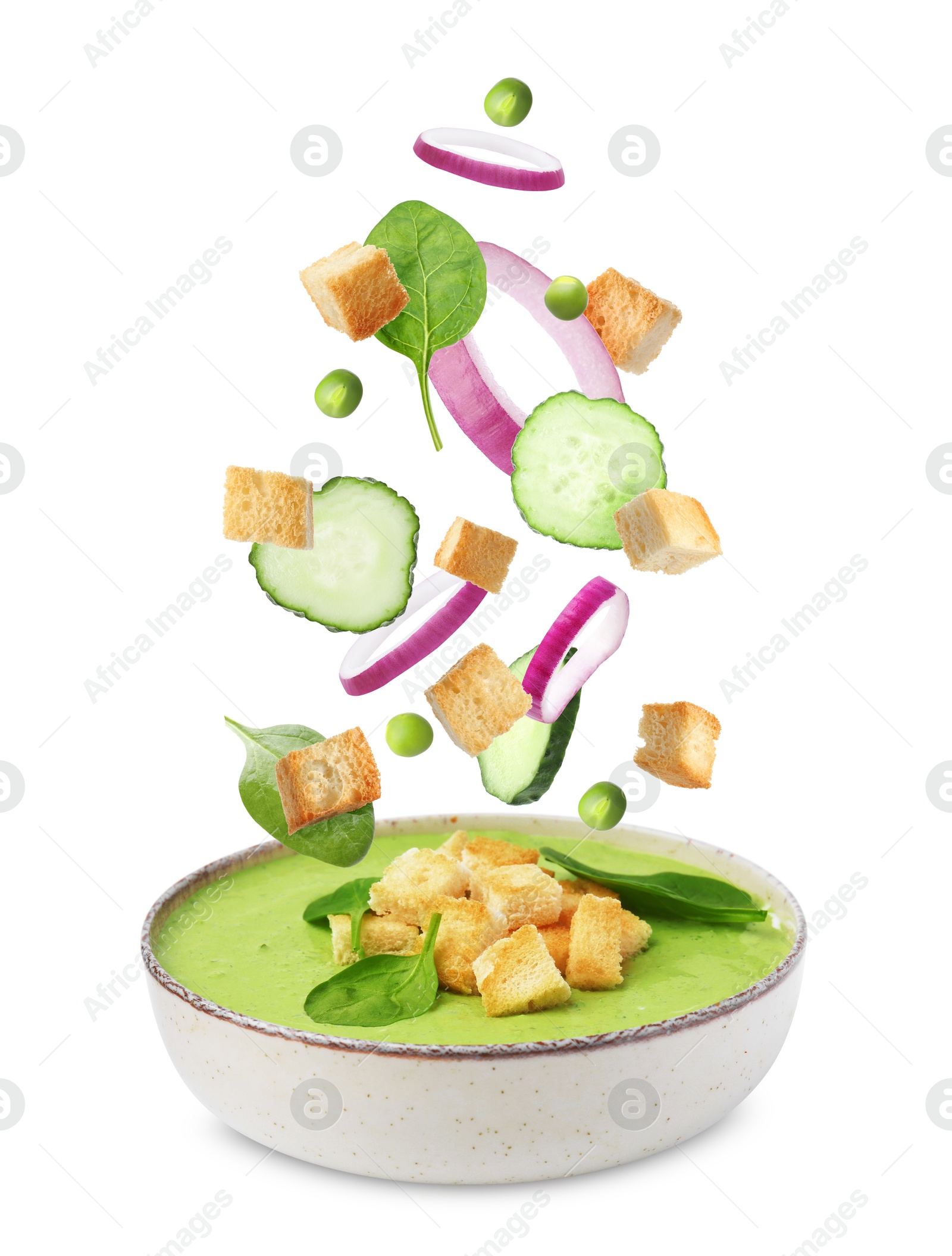 Croutons, onion rings, cucumber slices and green peas falling into bowl of soup against white background Image of Croutons, onion rings, cucumber slices and green peas falling into bowl of soup against white background