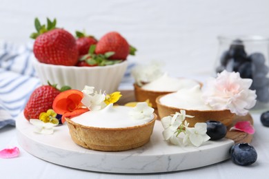 Photo of Delicious dessert. Tartlets with cream, berries and flowers on white tiled table, closeup