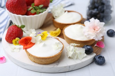Photo of Delicious dessert. Tartlets with cream, berries and flowers on white tiled table, closeup