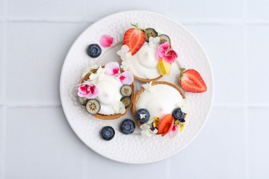 Photo of Delicious dessert. Tasty tartlets with cream, berries and flowers on white tiled table, top view