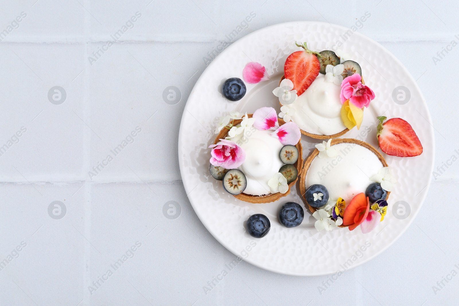 Photo of Delicious dessert. Tartlets with cream, berries and flowers on white tiled table, top view. Space for text