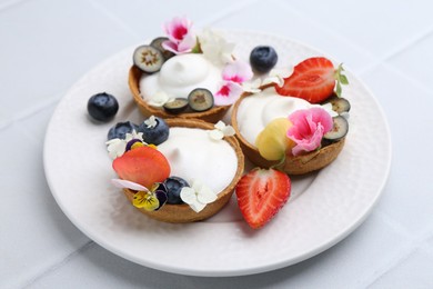 Photo of Delicious dessert. Tartlets with cream, berries and flowers on white tiled table, closeup