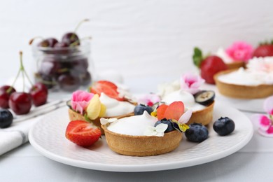 Photo of Delicious dessert. Tartlets with cream, berries and flowers on white tiled table, closeup