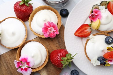 Photo of Delicious dessert. Tartlets with cream, berries and flowers on white tiled table, flat lay