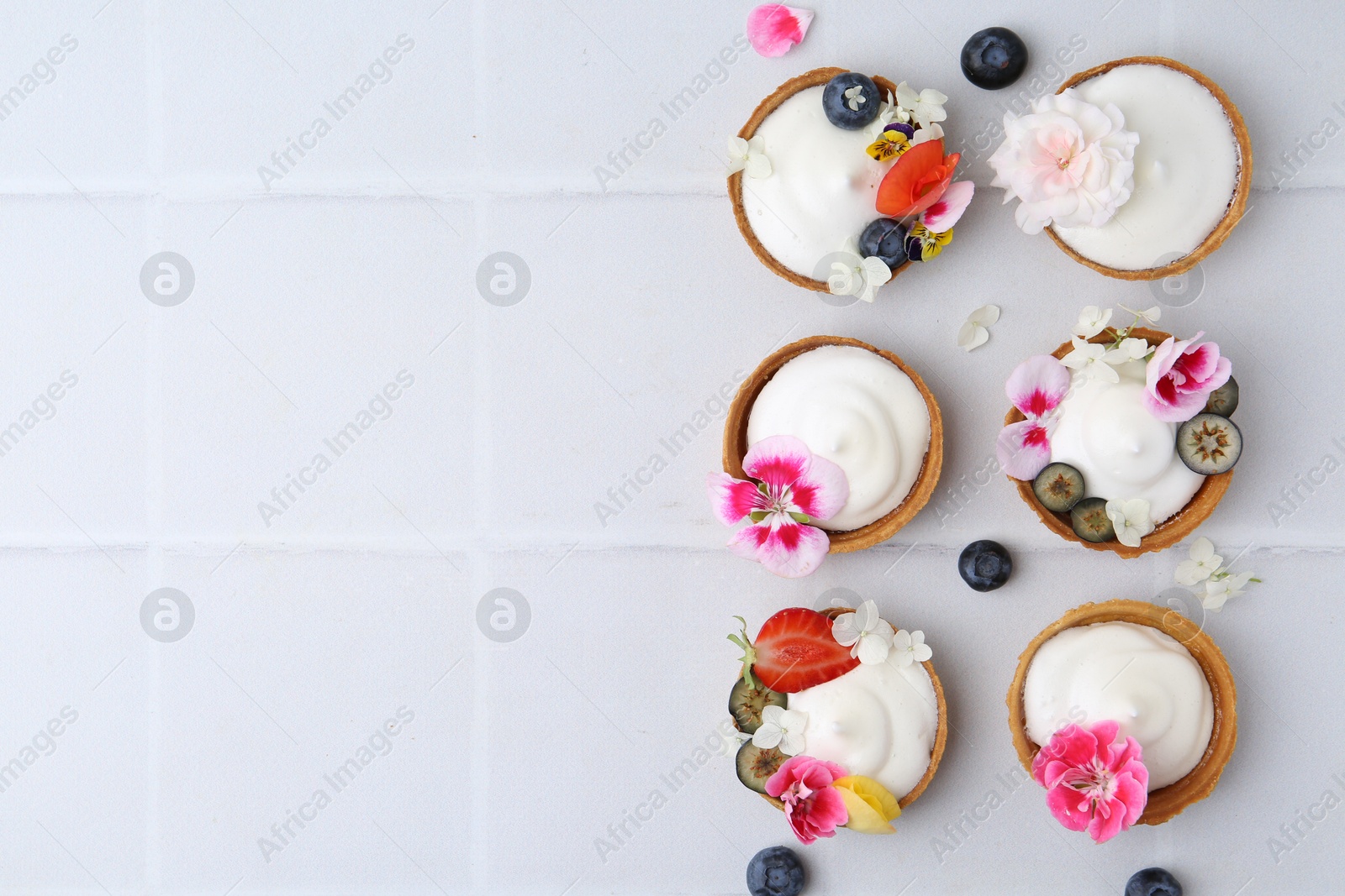Delicious dessert. Tartlets with cream, berries and flowers on white tiled table, flat lay. Space for text Photo of Delicious dessert. Tartlets with cream, berries and flowers on white tiled table, flat lay. Space for text