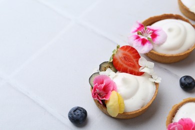 Photo of Delicious dessert. Tartlets with cream, berries and flowers on white tiled table, closeup. Space for text