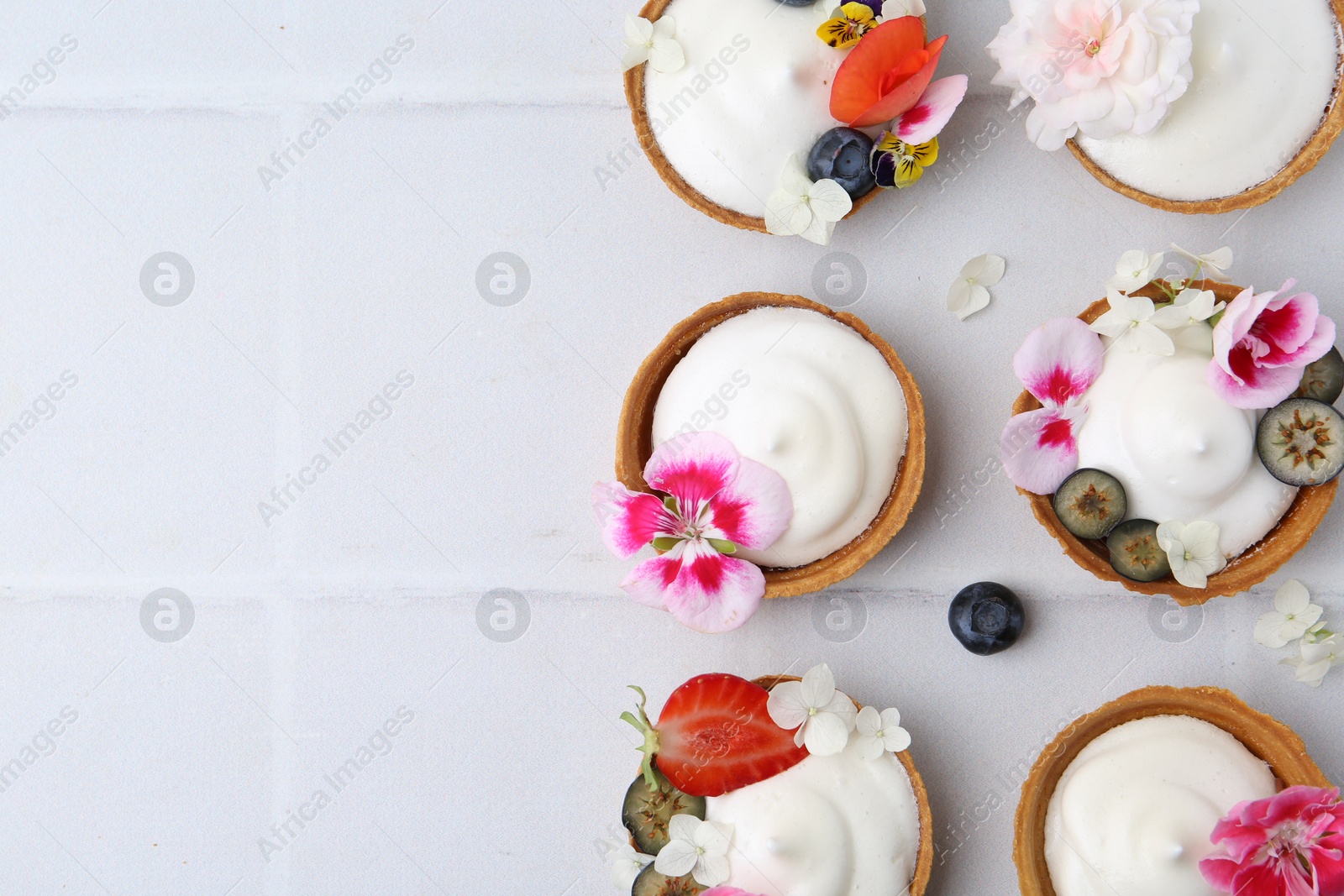 Delicious dessert. Tartlets with cream, berries and flowers on white tiled table, flat lay. Space for text Photo of Delicious dessert. Tartlets with cream, berries and flowers on white tiled table, flat lay. Space for text