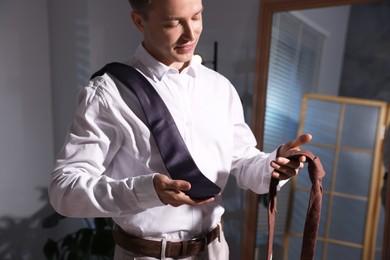 Photo of Handsome man choosing neckties while getting ready near mirror indoors