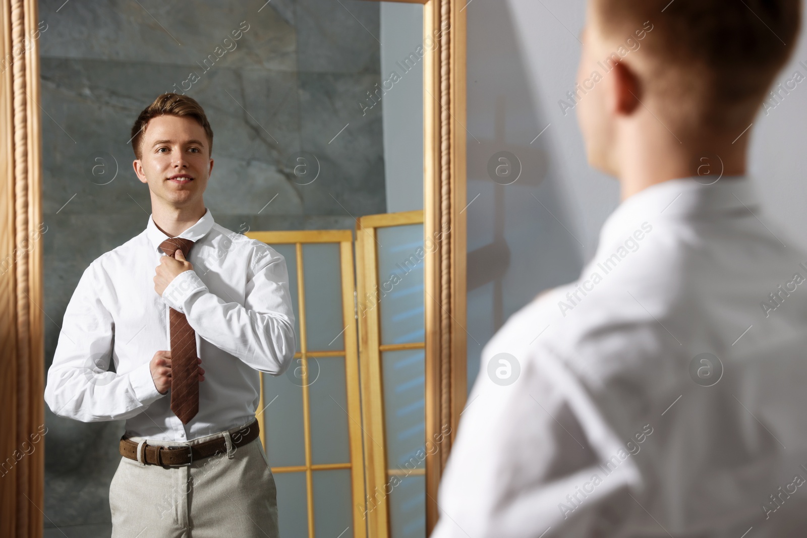 Handsome man adjusting necktie while getting ready near mirror indoors Photo of Handsome man adjusting necktie while getting ready near mirror indoors
