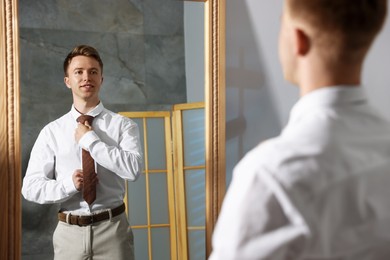 Photo of Handsome man adjusting necktie while getting ready near mirror indoors