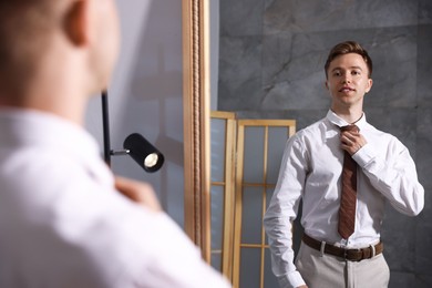 Photo of Handsome man adjusting necktie while getting ready near mirror indoors