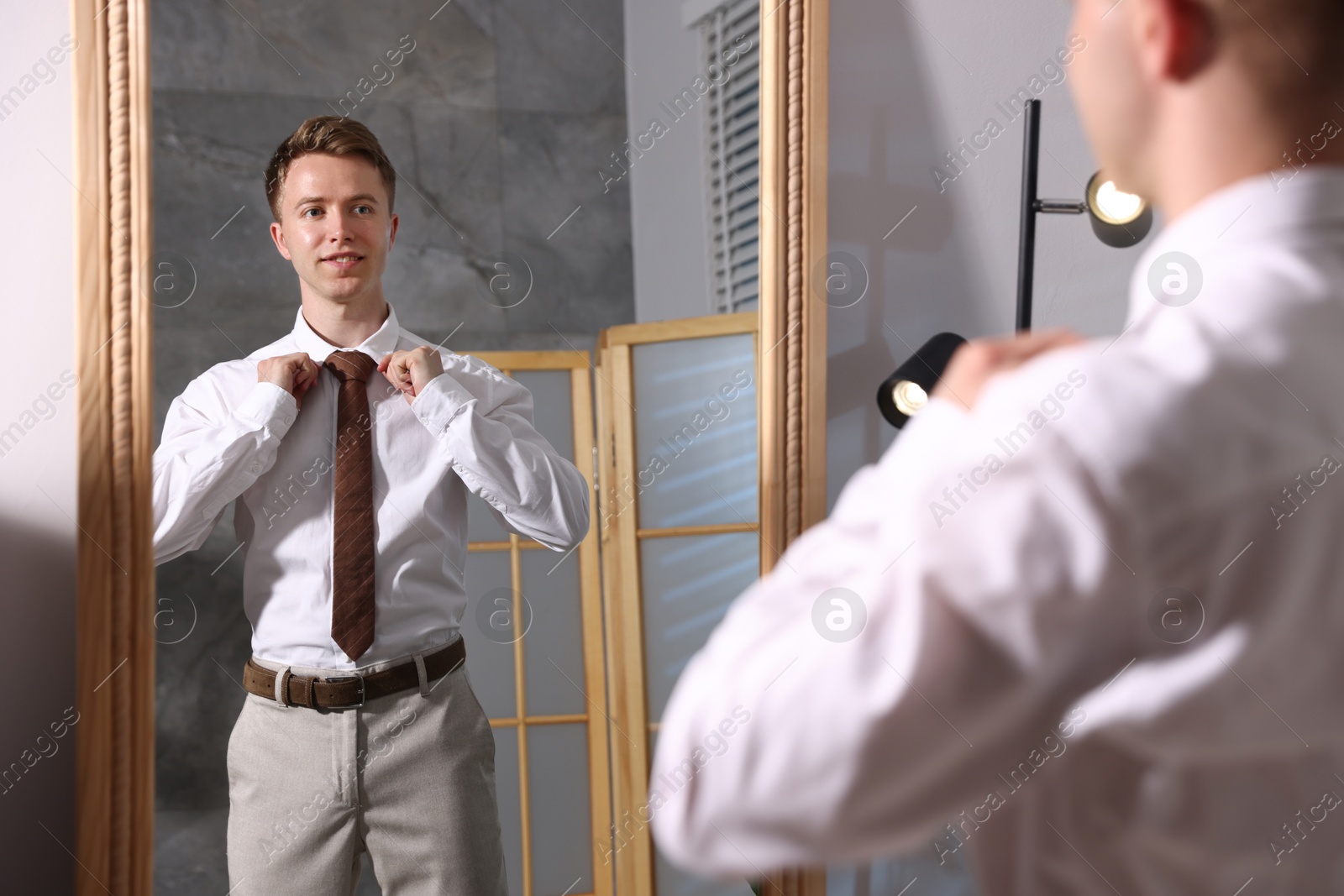 Handsome man adjusting necktie while getting ready near mirror indoors Photo of Handsome man adjusting necktie while getting ready near mirror indoors