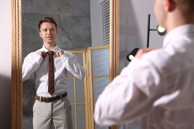 Photo of Handsome man adjusting necktie while getting ready near mirror indoors