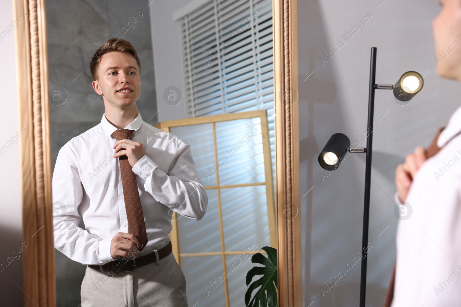 Handsome man adjusting necktie while getting ready near mirror indoors Photo of Handsome man adjusting necktie while getting ready near mirror indoors
