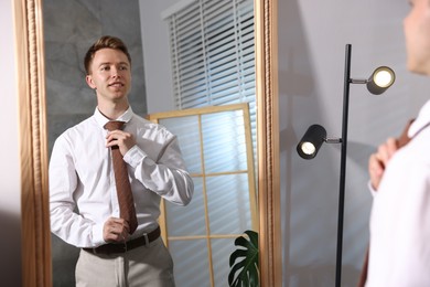Photo of Handsome man adjusting necktie while getting ready near mirror indoors