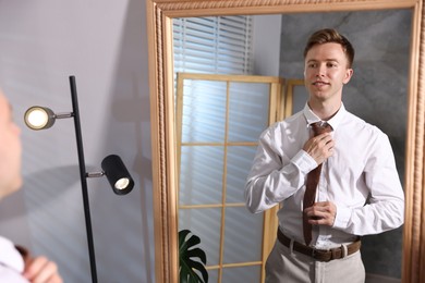Photo of Handsome man adjusting necktie while getting ready near mirror indoors
