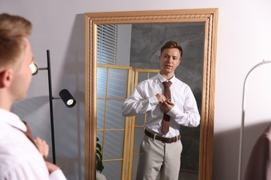 Photo of Man fastening tie clip to necktie while getting ready near mirror indoors