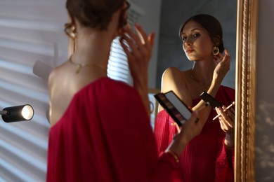 Photo of Beautiful woman with eyeshadow palette and brush getting ready near mirror indoors