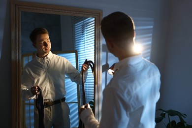 Photo of Man choosing neckties while getting ready near mirror indoors