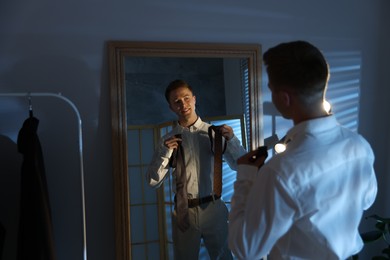 Photo of Man choosing neckties while getting ready near mirror indoors