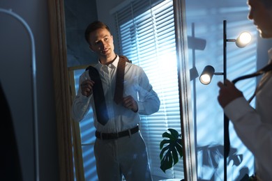 Photo of Man choosing neckties while getting ready near mirror indoors