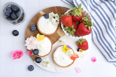 Photo of Delicious dessert. Tartlets with cream, berries and flowers on white tiled table, flat lay