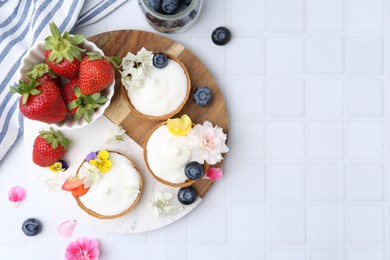 Photo of Delicious dessert. Tartlets with cream, berries and flowers on white tiled table, flat lay. Space for text