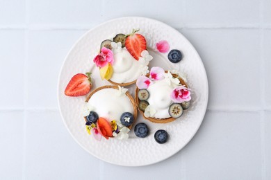 Photo of Delicious dessert. Tasty tartlets with cream, berries and flowers on white tiled table, top view