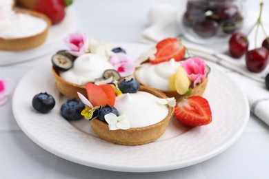 Photo of Delicious dessert. Tartlets with cream, berries and flowers on white tiled table, closeup