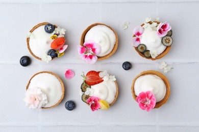 Photo of Delicious dessert. Tartlets with cream, berries and flowers on white tiled table, flat lay