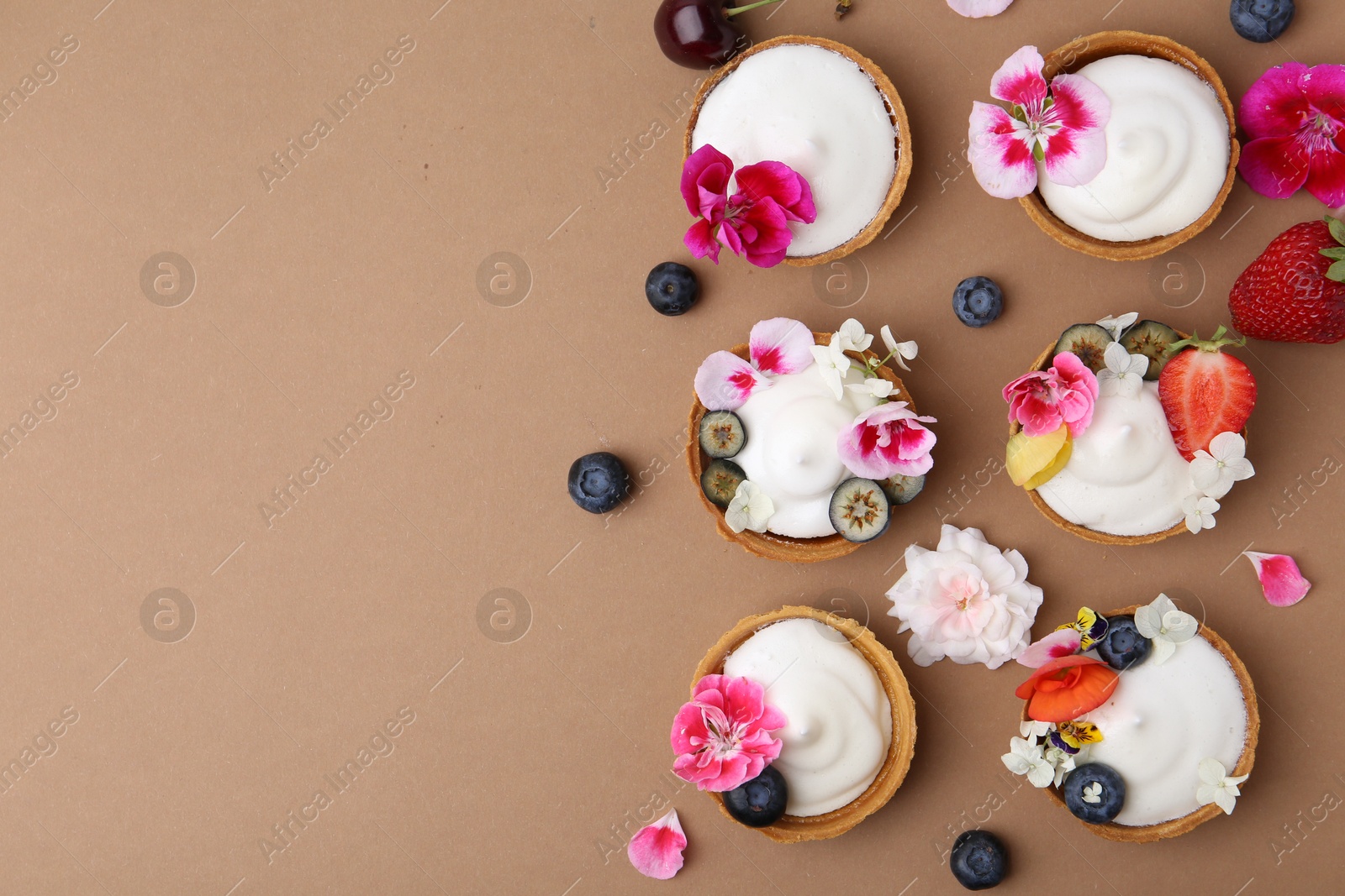 Delicious dessert. Tartlets with cream, berries and flowers on light brown background, flat lay. Space for text Photo of Delicious dessert. Tartlets with cream, berries and flowers on light brown background, flat lay. Space for text