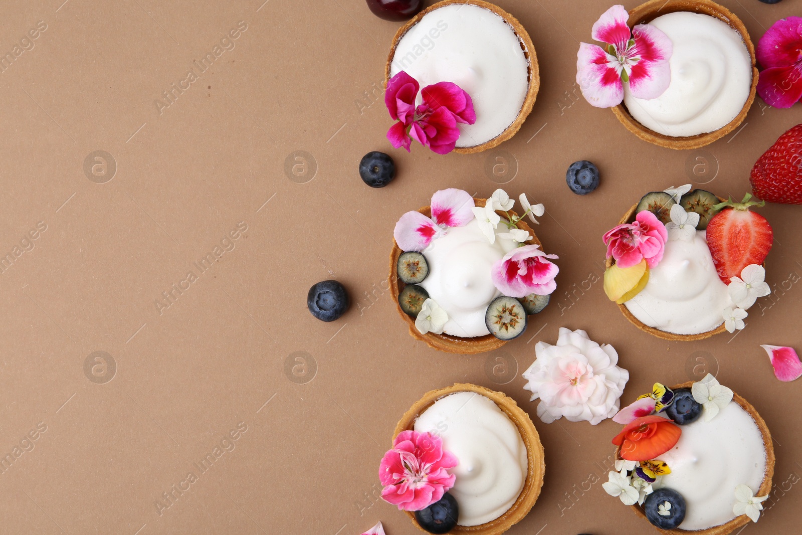 Delicious dessert. Tartlets with cream, berries and flowers on light brown background, flat lay. Space for text Photo of Delicious dessert. Tartlets with cream, berries and flowers on light brown background, flat lay. Space for text