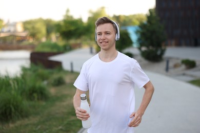 Photo of Smiling sportsman in headphones running with thermo bottle outdoors. Morning exercise