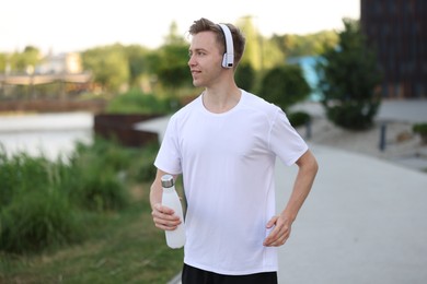 Photo of Smiling sportsman in headphones running with thermo bottle outdoors. Morning exercise