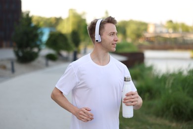 Photo of Smiling sportsman in headphones running with thermo bottle outdoors. Morning exercise