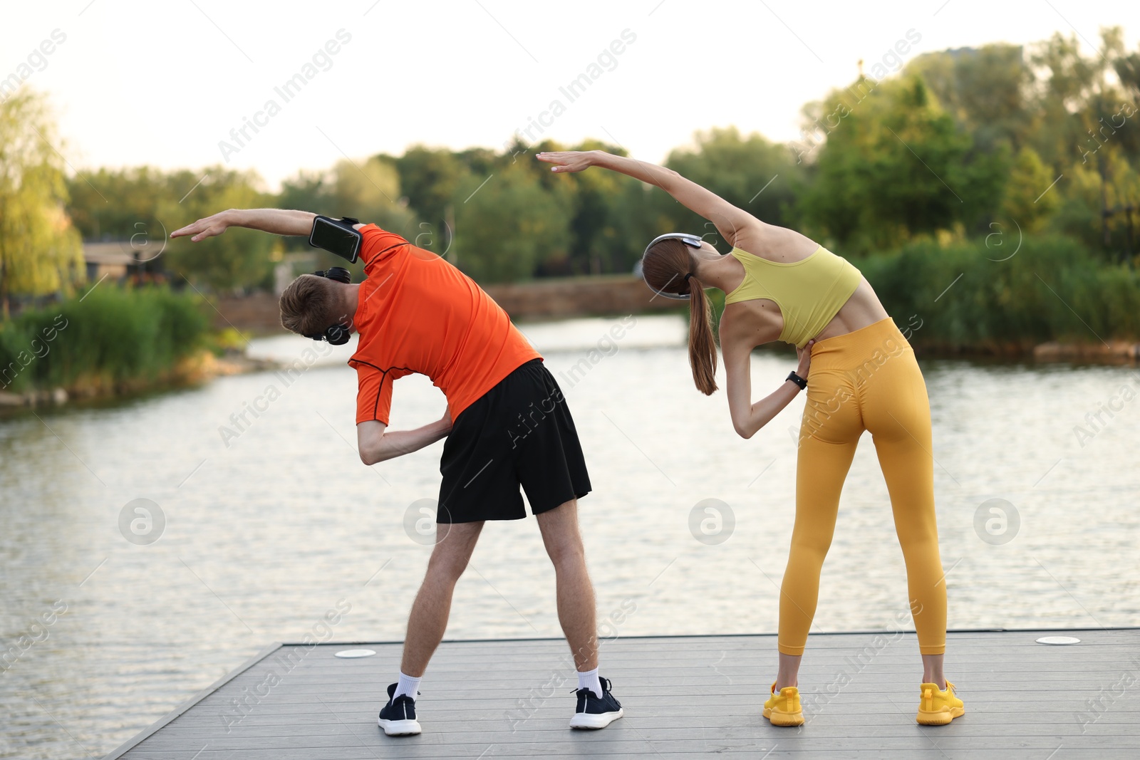 Couple in headphones doing morning exercise on pier near river, back view Photo of Couple in headphones doing morning exercise on pier near river, back view