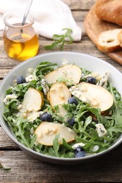 Photo of Tasty salad with arugula, pear, blue cheese, peanuts and blueberries in bowl on wooden table, closeup