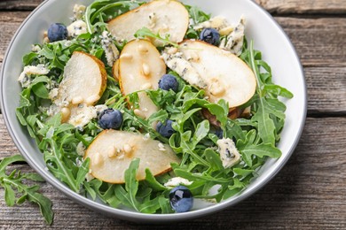 Photo of Tasty salad with arugula, pear, blue cheese, peanuts and blueberries in bowl on wooden table, above view