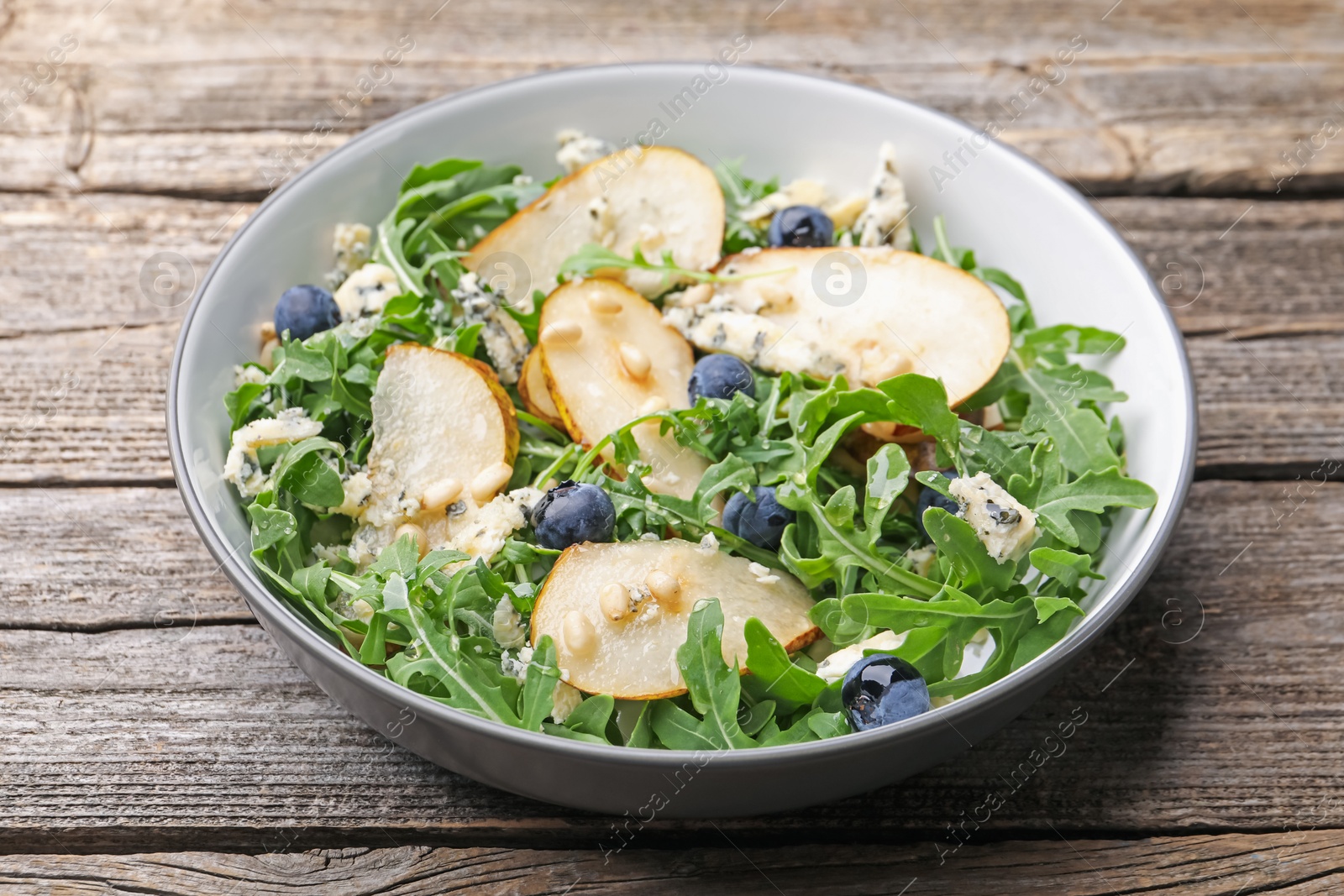 Photo of Tasty salad with arugula, pear, blue cheese, peanuts and blueberries in bowl on wooden table, closeup
