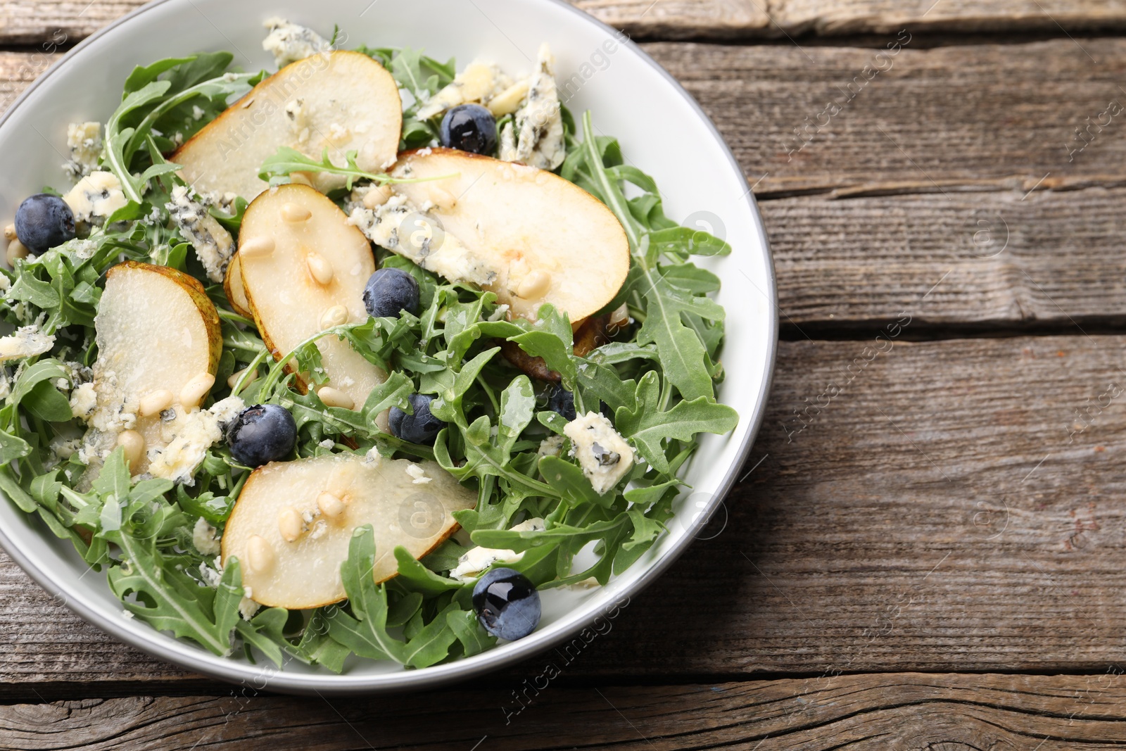 Photo of Tasty salad with arugula, pear, blue cheese, peanuts and blueberries in bowl on wooden table, above view. Space for text