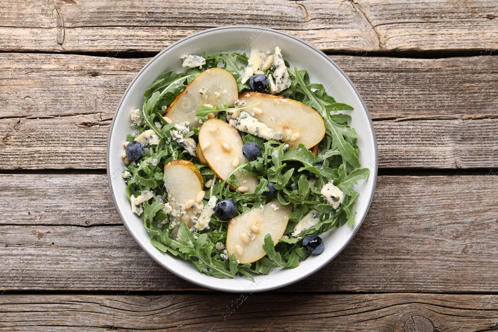 Tasty salad with arugula, pear, blue cheese, peanuts and blueberries in bowl on wooden table, top view Photo of Tasty salad with arugula, pear, blue cheese, peanuts and blueberries in bowl on wooden table, top view