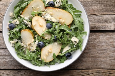 Photo of Tasty salad with arugula, pear, blue cheese, peanuts and blueberries in bowl on wooden table, top view